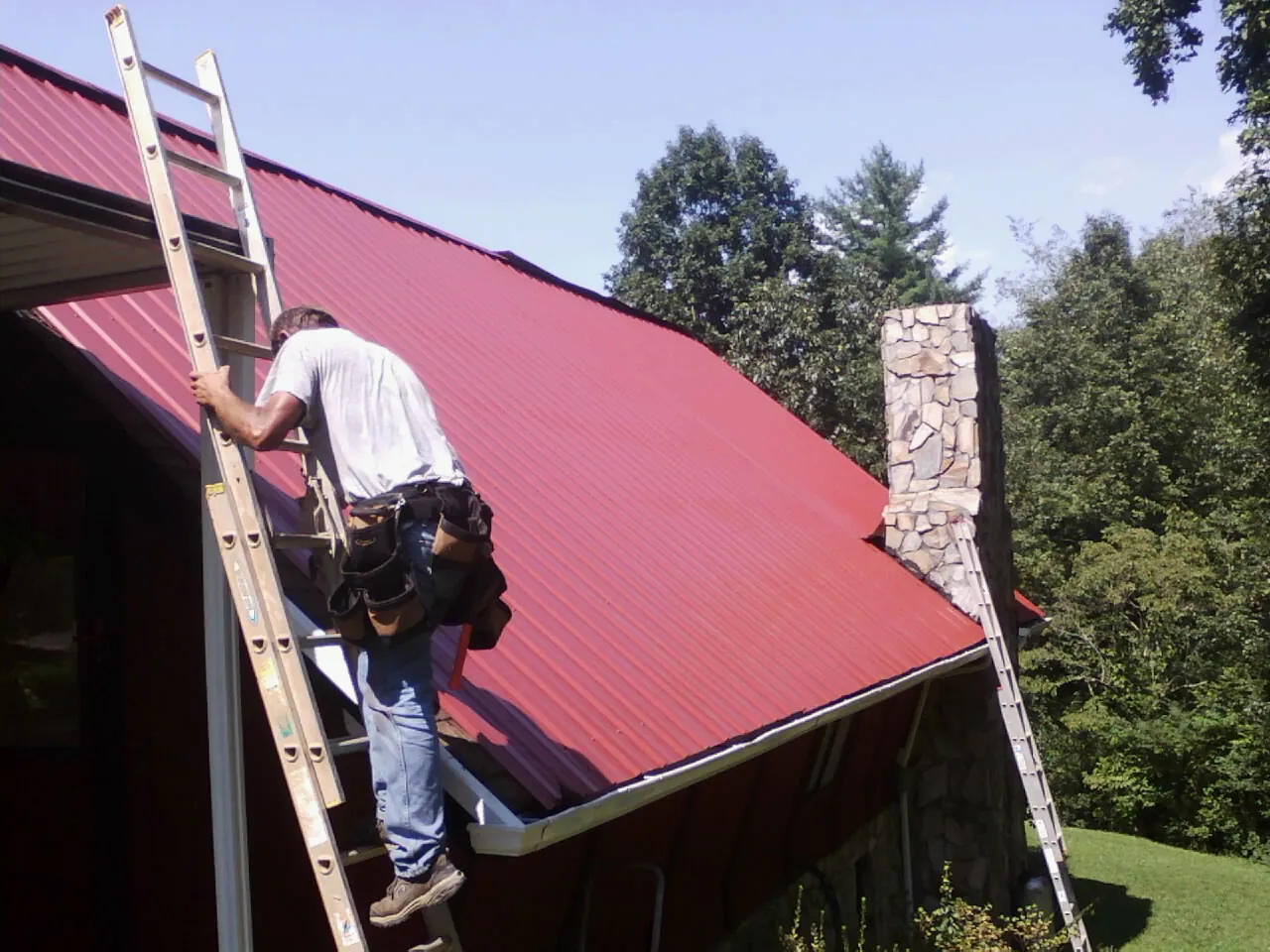Roofer on a ladder beside a red metal roof and stone chimney — Soffit & Fascia Repair by Roofing Brothers in Foresthill